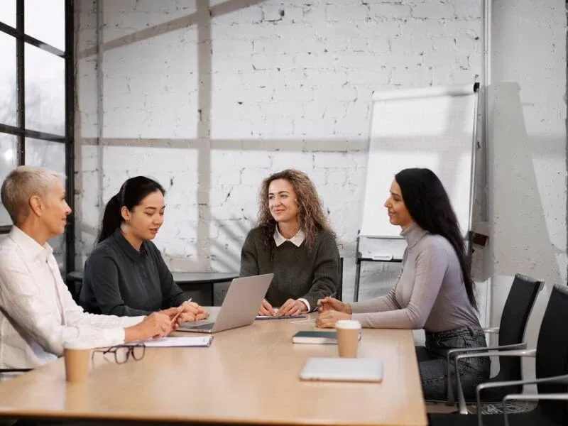 A small group of professionals seated around a table in a bright meeting room, discussing notes beside a flipchart